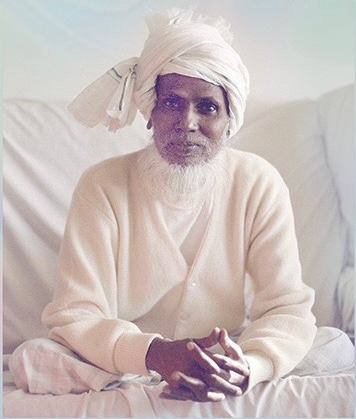 Portrait of Bawa Muhaiyaddeen seated with folded hands in a contemplative posture.