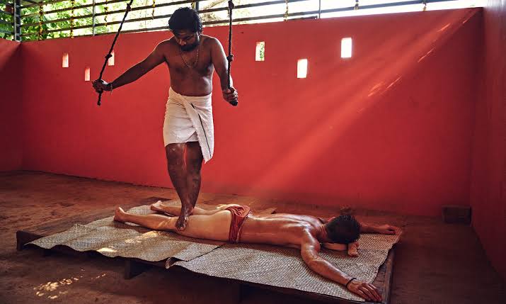 Practitioner performing traditional Uzhichil foot-pressure massage while holding suspended ropes for balance in a Kerala Kalari.