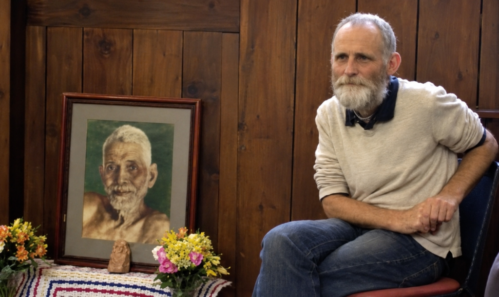 Michael James seated beside a framed portrait of Ramana Maharshi during an earlier period of his study and practice of Ramana’s teachings.