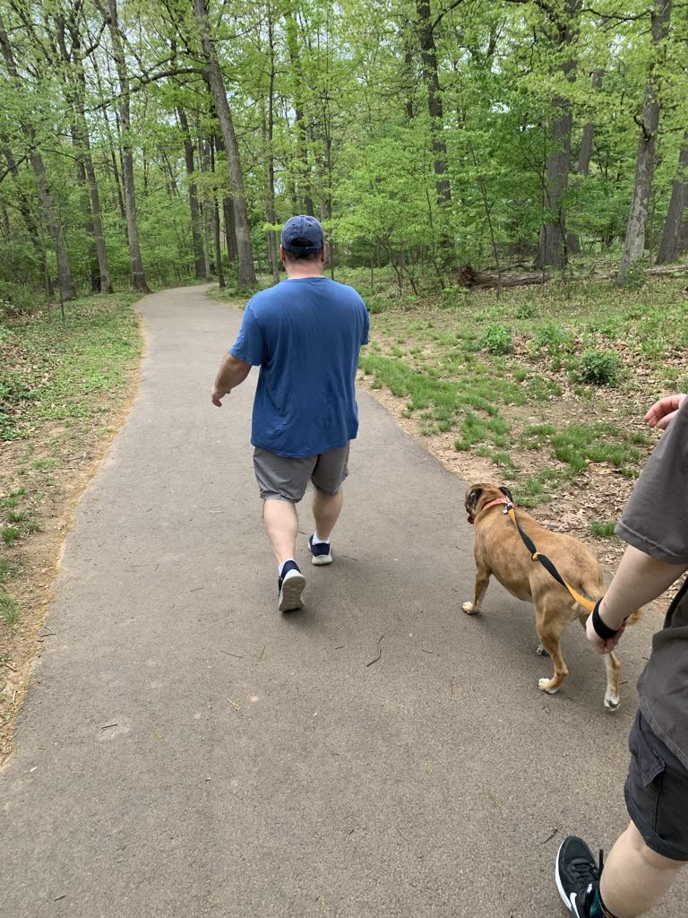 Mark V. Wiley walking away from the camera along a paved woodland trail, with his dog and a companion following behind.