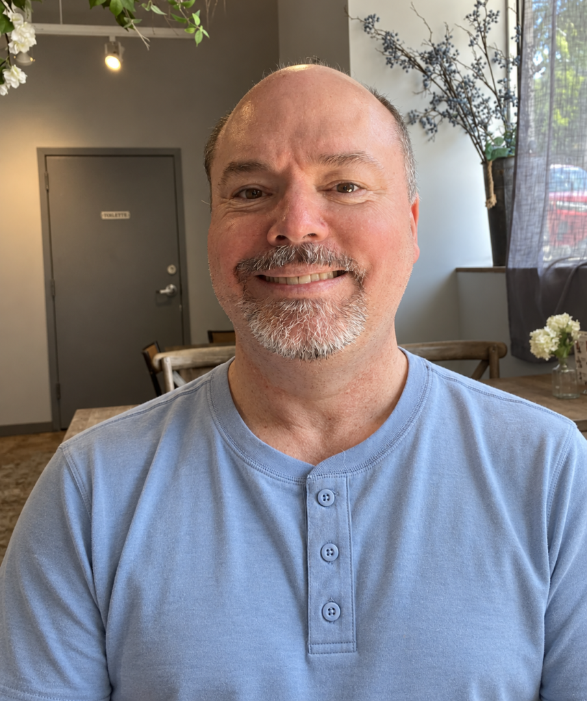 Wiley smiling in a light blue shirt, seated in a café with plants and natural window light.